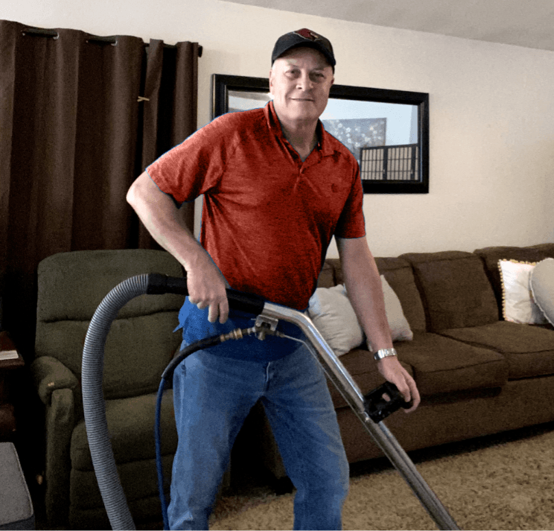 A smiling man in a red shirt and baseball cap uses a carpet cleaner in a living room.